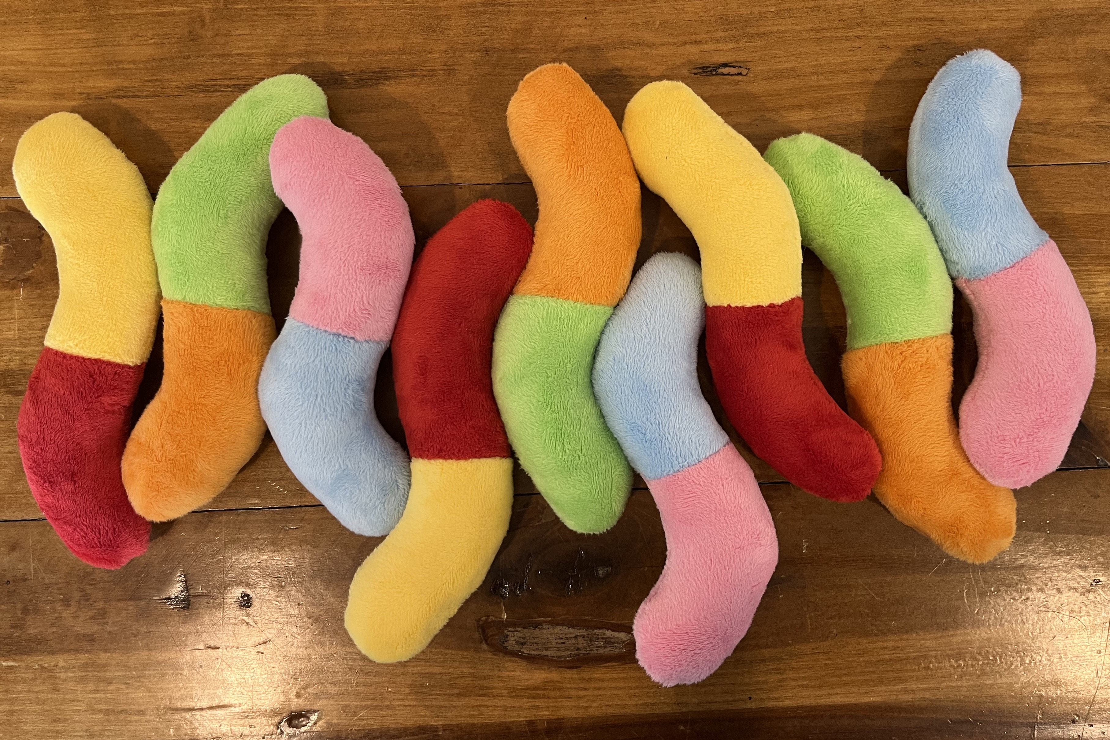 A pile of plush gummy worms in green/orange, red/yellow, and pink/blue laying on a wooden table
