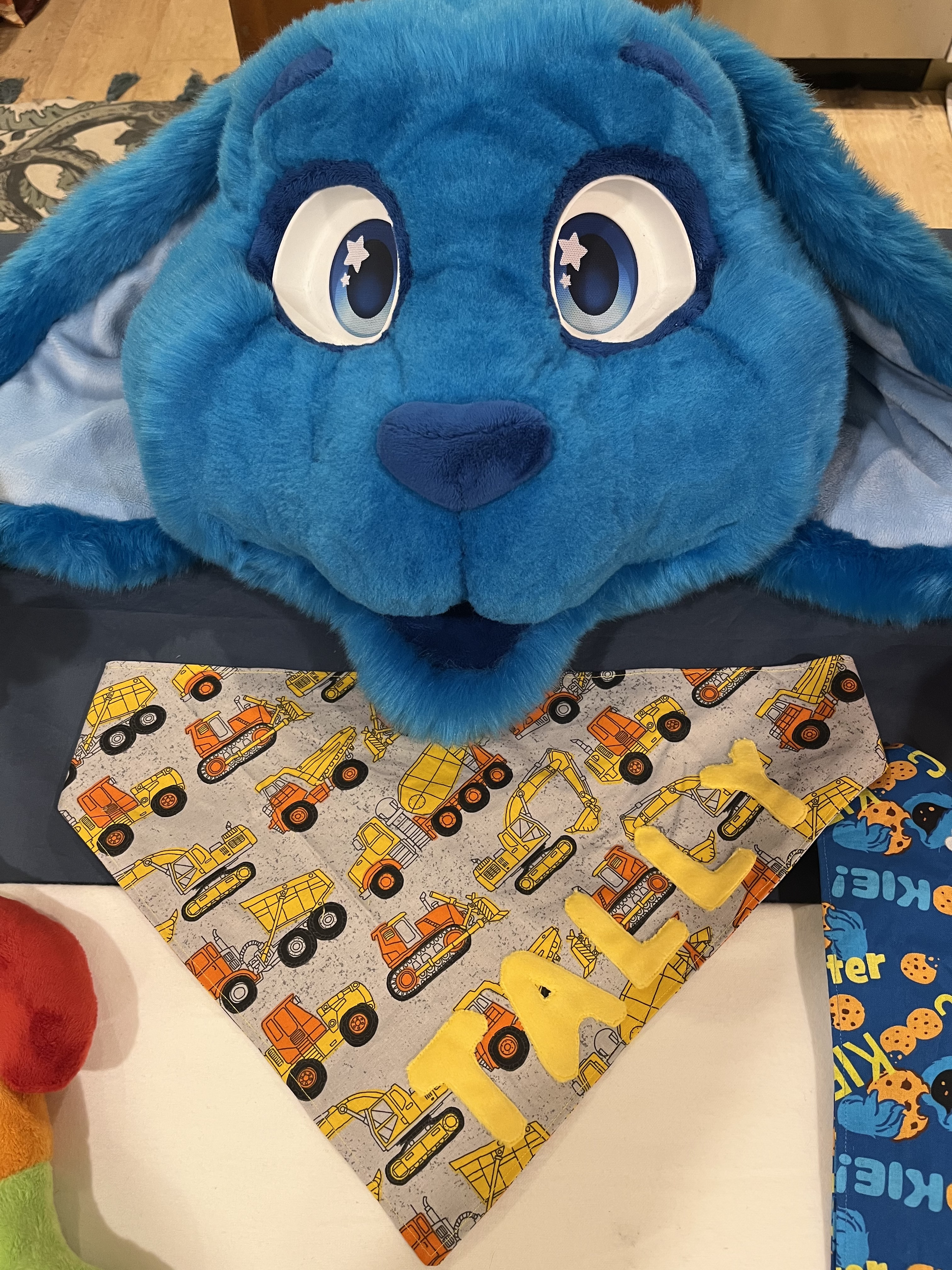 A blue rabbit fursuit head smiling next to a construction-print bandana with Tally appliqued on it