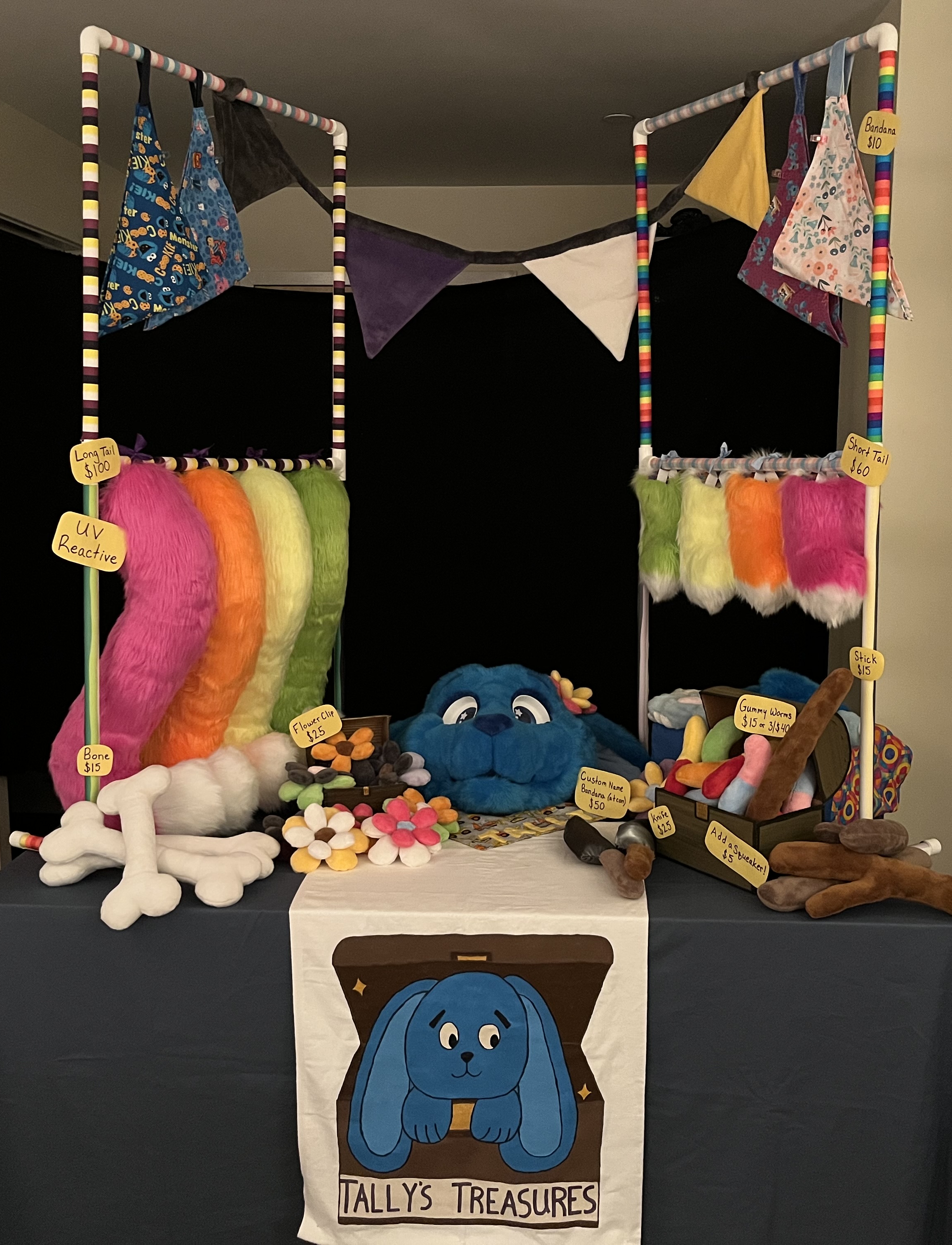 My booth setup. A table with a blue tablecloth, a banner reading Tally's Treasures, two stands with neon fursuit tails and bandanas hanging from them, and plush sticks, bones, knives, flowers, and gummy worms displayed on the table.