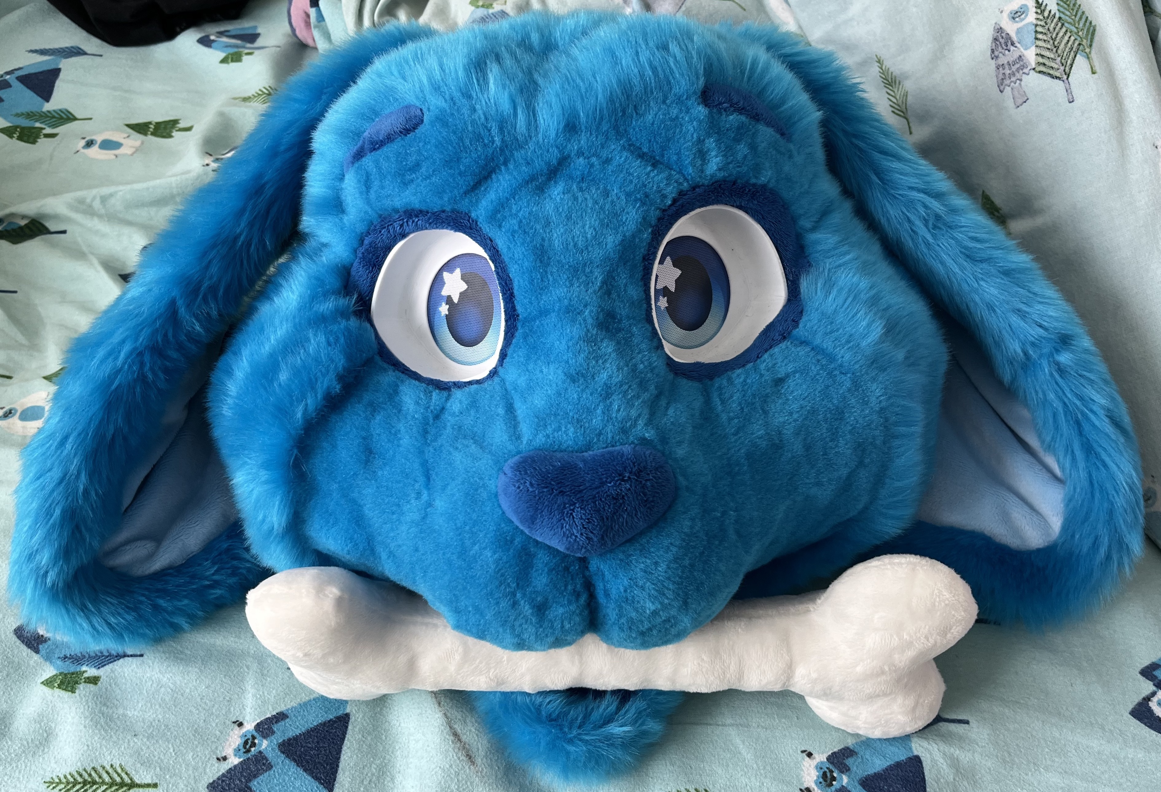 A blue rabbit fursuit head holding an off-white plush dog bone in its mouth