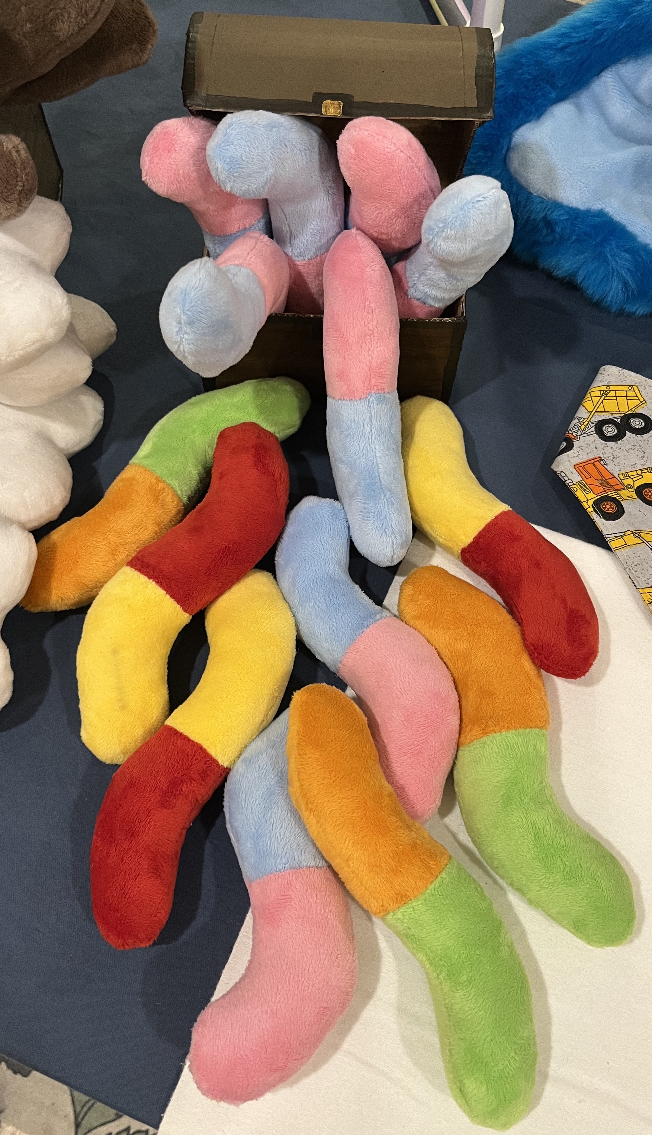 A pile of plush gummy worms in blue/pink, red/yellow, and orange/green spilling out of a small treasure chest onto a table