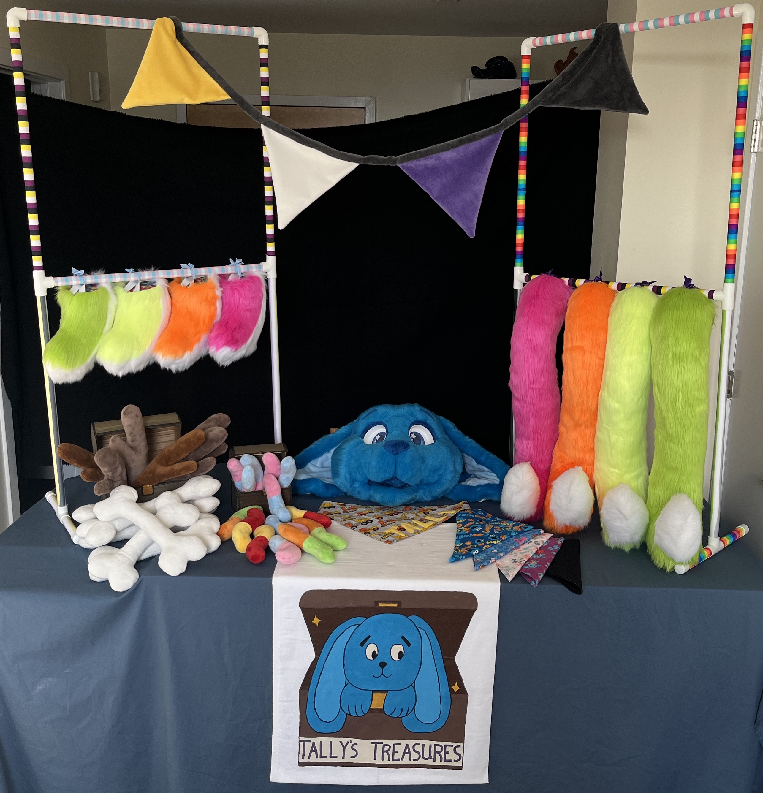 My booth setup in my living room. A table with a blue tablecloth, a banner reading Tally's Treasures, two stands with neon fursuit tails hanging from them, and bandanas, plush sticks, plush bones, and plush gummy worms laying on the table.