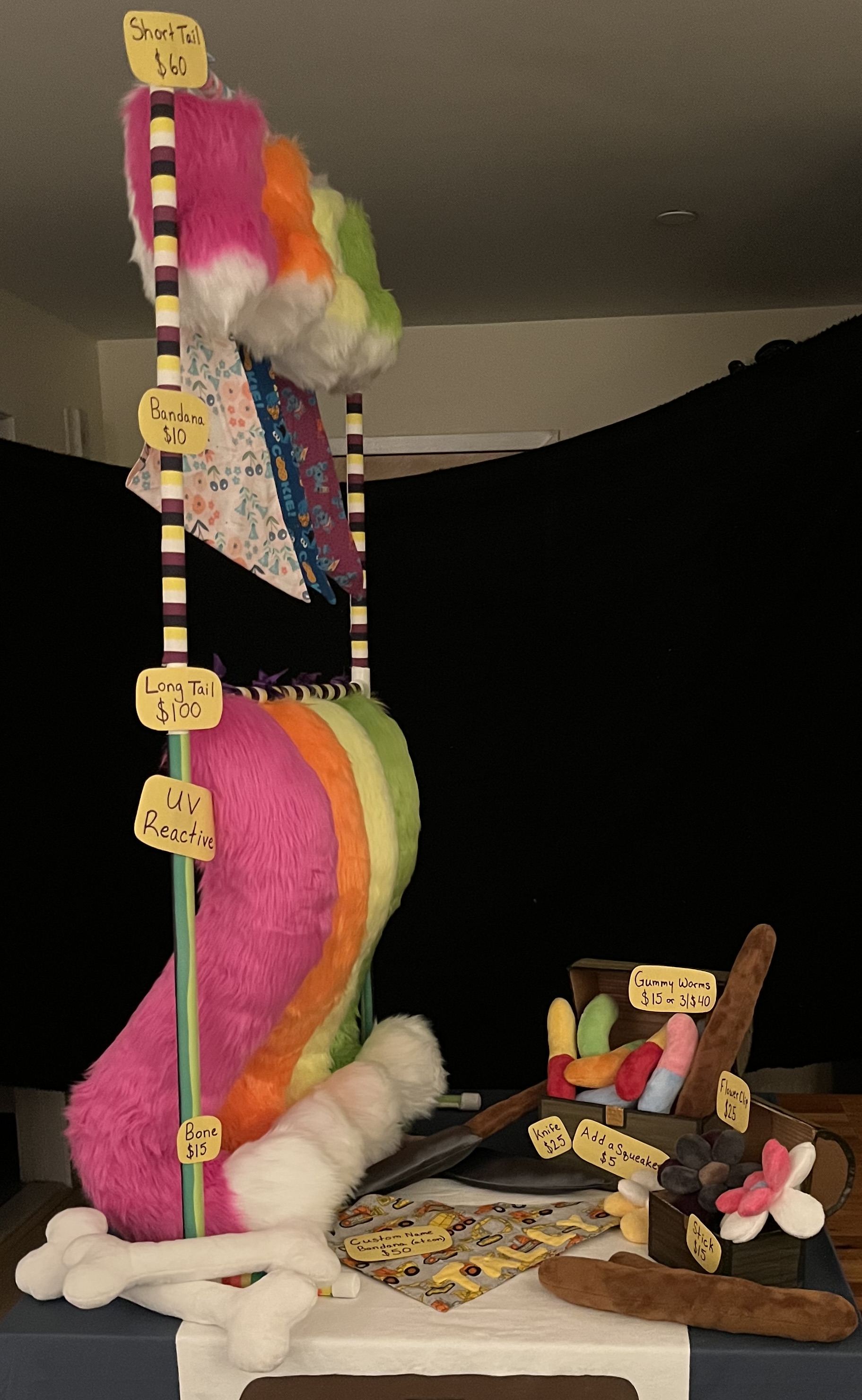 A small table with a blue tablecloth, a stand with neon fursuit tails and bandanas hanging from it, and plush sticks, bones, flowers, knives, and gummy worms displayed on the table.
